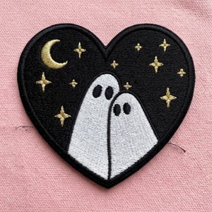 May include: A heart-shaped embroidered patch featuring a black background with two white ghosts standing side by side. The design includes a golden crescent moon and several golden four-pointed stars scattered across the dark night sky.