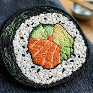 May include: A circular embroidered patch featuring a sushi roll design. The center displays orange salmon, light green avocado, and a small cluster of bright green wasabi. These are surrounded by white rice and a dark green seaweed outer border.