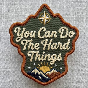 May include: An embroidered patch with a dark green background and a copper-colored border. It features a white compass rose at the top, the text "You Can Do The Hard Things" in white, and a yellow sun rising over blue and brown mountain peaks.