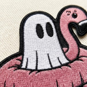 May include: An embroidered patch featuring a white ghost with black oval eyes sitting inside a pink flamingo pool float. The design is outlined in thick black thread, showcasing detailed stitching throughout the ghost and the textured pink flamingo body.