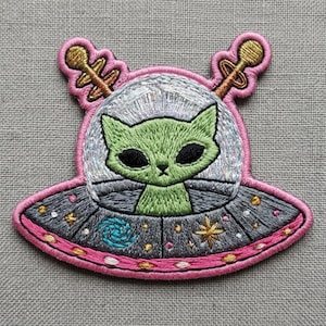 May include: An embroidered patch featuring a green cat with large black eyes inside a silver and grey UFO. The spacecraft has a pink border, two gold antennas, and is decorated with small gold stars and a blue spiral galaxy design.