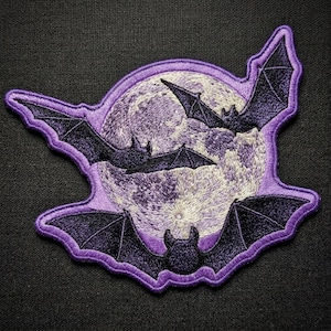 May include: An embroidered patch featuring a large, detailed full moon in shades of cream and light purple, surrounded by three dark purple bats with outstretched wings. The entire design is outlined with a thick, vibrant purple border.