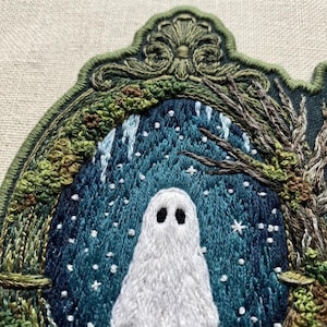 May include: An embroidered patch featuring a white ghost centered within a dark blue, snowy forest scene. The scene is framed by an ornate, olive green stitched border with intricate scrollwork at the top and textured foliage on the sides.
