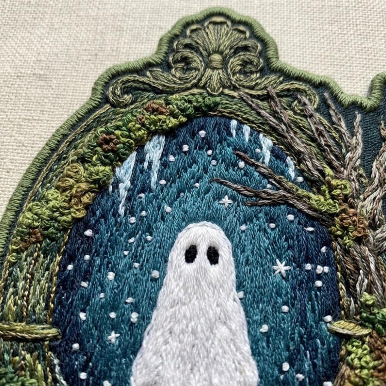 May include: An embroidered patch featuring a white ghost centered within a dark blue, snowy forest scene. The scene is framed by an ornate, olive green stitched border with intricate scrollwork at the top and textured foliage on the sides.