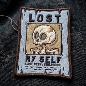 May include: A rectangular embroidered patch with a light blue background and a dark brown border. It features a beige square containing a cartoon skull and skeletal torso. The text reads LOST MY SELF and LAST SEEN: CHILDHOOD in dark brown.