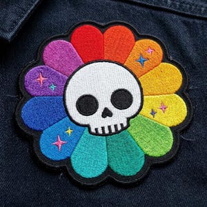 May include: An embroidered patch featuring a central white skull with black eye sockets and nose, surrounded by twelve rounded petals in a rainbow gradient of red, orange, yellow, green, blue, and purple, accented with small embroidered stars.