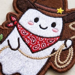 May include: An embroidered patch featuring a white ghost character wearing a brown cowboy hat with a yellow star, a red paisley bandana, and holding a tan rope lasso. The ghost has black eyes, a small smile, and pink cheeks, with brown stitching.