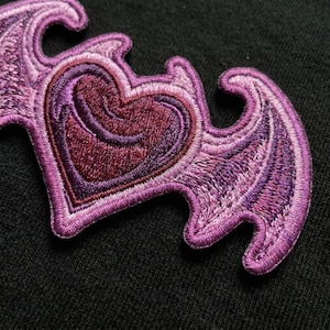 May include: An embroidered patch featuring a central heart shape in shades of deep purple and magenta, flanked by two stylized bat wings in lighter lavender and violet tones. The design is rendered with dense thread work and a raised texture.