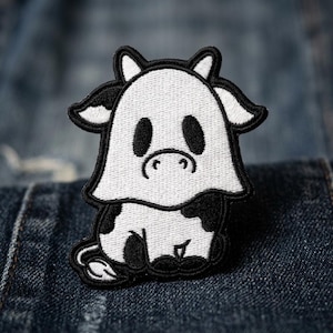 May include: An embroidered patch featuring a cartoon cow wearing a white ghost sheet. The design is rendered in black and white thread with a thick black border, depicting the cow sitting down with its tail visible at the bottom left corner.