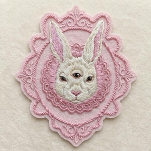 May include: An embroidered patch featuring a three-eyed rabbit head centered within a pink scalloped frame. The rabbit has cream-colored fur, pink-tinted ears, and brown eyes. The frame is decorated with intricate pink rope and lace-like stitching.