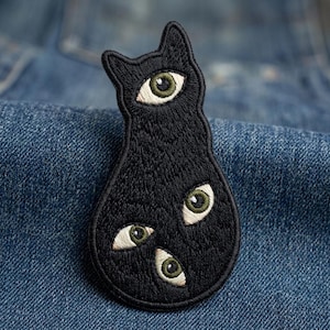 May include: An embroidered patch in the shape of a black cat silhouette. The cat figure features three realistic human-like eyes with green irises and white sclera, with one eye positioned on the head and two eyes placed on the lower body section.