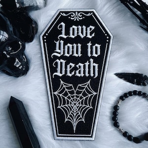 May include: A black coffin-shaped embroidered patch featuring white stitched borders and the text "Love You to Death" in a gothic font. Below the text is a white embroidered spiderweb design shaped like a heart on a black fabric background.