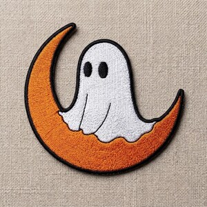 May include: An embroidered patch featuring a white ghost with two black oval eyes sitting on a bright orange crescent moon. The entire design is outlined in a thick black thread, creating a clear contrast between the ghost and the moon shape.