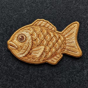 May include: An embroidered patch in the shape of a fish, featuring golden-brown thread with detailed stitching to represent scales, fins, and a circular eye. The design is rendered in a monochromatic palette of warm tan and brown tones.