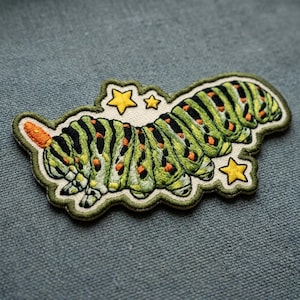 May include: An embroidered patch featuring a green caterpillar with black stripes and orange spots. The caterpillar has an orange horn on its head and is surrounded by three yellow stars on a cream-colored background with a dark green border.