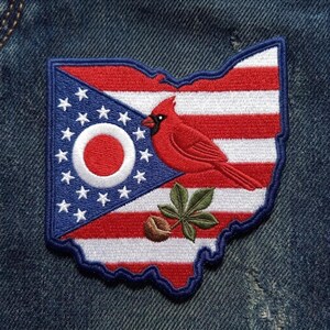 May include: An embroidered patch shaped like the state of Ohio featuring a red cardinal bird and a buckeye nut with leaves. The design incorporates the American flag pattern with red and white stripes, a blue triangle, white stars, and a white circle.