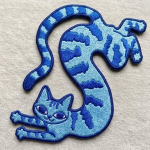 May include: An embroidered patch in the shape of a cat with a long, curved body. The cat is light blue with dark blue stripes, featuring a smiling face, pointed ears, and outstretched front paws. The entire design is outlined in dark blue thread.