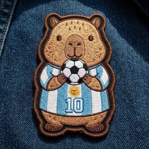 May include: An embroidered patch featuring a brown capybara wearing a light blue and white striped soccer jersey with the number 10 and a sun emblem on the chest. The capybara is holding a black and white soccer ball with both paws.