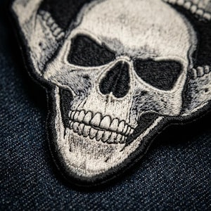 May include: An embroidered patch featuring a detailed human skull design in shades of white, gray, and black. The skull is depicted with deep eye sockets, a prominent nasal cavity, and visible teeth, rendered with intricate thread work and stitching.