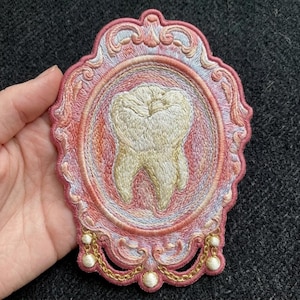 May include: An embroidered patch featuring a central cream-colored tooth motif enclosed within an ornate, oval-shaped frame. The frame is stitched in shades of pink, peach, and light blue, accented with gold-colored thread chains and small white beads.
