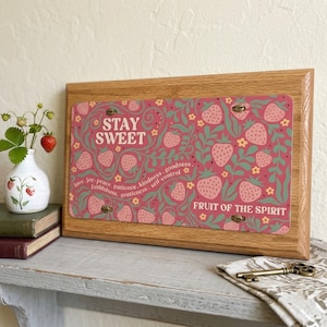 May include: A rectangular wooden plaque featuring a pink background with a repeating pattern of red strawberries, green leaves, and small yellow flowers. The text reads STAY SWEET and FRUIT OF THE SPIRIT with a list of virtues in white script.