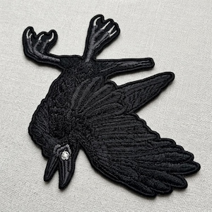 May include: An embroidered iron-on patch featuring a black raven in a downward-facing position with its wings spread wide. The bird has detailed black thread stitching throughout its feathers and a single small, clear rhinestone set as its eye.