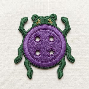 May include: An embroidered patch shaped like a beetle with a purple circular button body. The button features four holes, one of which is star-shaped. The beetle has dark green legs and a head with two small gold embroidered stars on top.