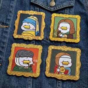 May include: Four square embroidered patches featuring cartoon ducks dressed as famous historical figures, each enclosed in a gold-colored ornate frame. The patches are arranged in a grid pattern on a dark blue denim fabric background.