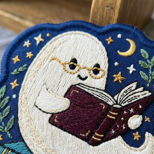 May include: An embroidered patch featuring a white ghost wearing gold-rimmed glasses and reading a dark maroon book. The background is deep blue with yellow stars, a crescent moon, and green leafy sprigs stitched along the edges of the fabric.