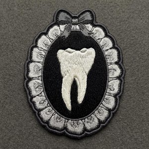 May include: An oval embroidered patch featuring a white tooth centered on a black background. The design is framed by a scalloped white border and topped with a small grey bow. The entire piece is stitched with detailed thread work.