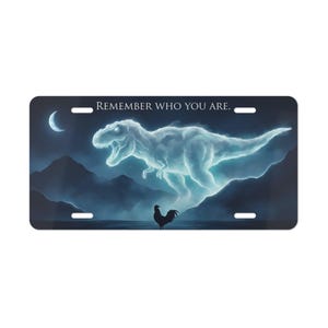 May include: License plate featuring a surreal illustration. A glowing, cloud-like Tyrannosaurus Rex dominates the scene, with a rooster below. The background depicts a moonlit landscape with mountains and water. The text "REMEMBER WHO YOU ARE" is at the top.