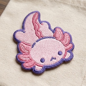 May include: An embroidered patch featuring a light pink axolotl with dark purple outlines. The axolotl has a rounded head, two small dark purple eyes, a simple mouth, and prominent feathery pink gills on both sides of its head.