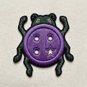 May include: An embroidered patch shaped like a beetle with a large purple button as its body. The button features four holes, one of which is star-shaped. The beetle has a dark green head with two small gold embroidered stars and six dark green legs.