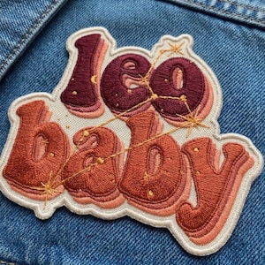 May include: An embroidered patch featuring the word Leo in dark maroon and baby in burnt orange, both in a bubble font. A gold thread constellation pattern overlays the letters, with small starburst accents on a cream-colored fabric border.