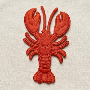 May include: An embroidered iron-on patch in the shape of a lobster. The lobster is a vibrant orange-red color with detailed stitching that defines its large claws, segmented body, multiple legs, and wide tail fan against a plain fabric background.