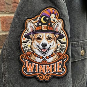 May include: An embroidered patch on a dark gray fabric featuring a Corgi wearing a purple and orange witch hat with a crescent moon. The design includes spiderwebs, bats, small pumpkins, a skull bead necklace, and the text WINNIE in orange.