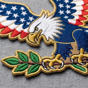 May include: An embroidered patch featuring a bald eagle with white head feathers, a yellow beak, and wings patterned with red, white, and blue stripes and white stars. The eagle holds a green olive branch with yellow talons, outlined in gold thread.