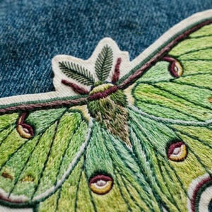 以下が含まれることがあります： 明るい緑とライムグリーンの色合いで描かれた、オオミズアオの刺繍ワッペン。蛾には濃い栗色の翅脈、羽毛のような触角、そして翅には黄色と栗色の中心を持つ円形の目玉模様が施されています。