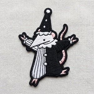 May include: An embroidered patch featuring a white mouse dressed as a jester in a black and white outfit. The mouse wears a black pointed hat with white dots, a white ruffled collar, and a black and white striped tunic with two white buttons.