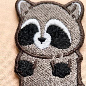 May include: An embroidered patch featuring a stylized raccoon with light brown fur, a white face mask, black eye patches, a small black nose, and two small black paws. The design is outlined with a dark brown border and has a textured fabric finish.
