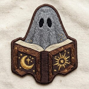 May include: An embroidered patch featuring a grey ghost figure with two black oval eyes positioned above an open brown book. The book cover displays a gold crescent moon and stars on the left and a gold sunburst design on the right.