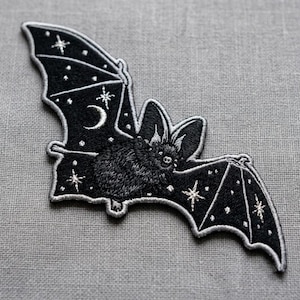 May include: A black embroidered patch in the shape of a bat with spread wings. The design features white stitched details including a crescent moon and multiple four-pointed stars scattered across the wings, with a textured body and ears.