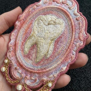May include: An embroidered patch featuring a central cream-colored tooth motif encased within an ornate, scalloped oval frame in shades of pink, peach, and lavender. The border is accented with metallic gold thread and small white beads.