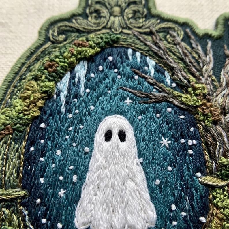 May include: A detailed embroidered patch featuring a white ghost with black eyes centered against a dark teal background. The scene is framed by an ornate green border with stitched tree branches, mossy textures, and small white star accents.