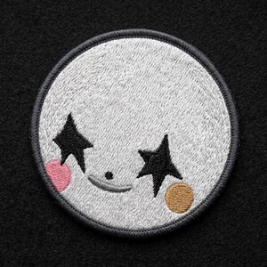 May include: A circular embroidered patch featuring a light gray face with two black star-shaped eyes, a small black dot for a nose, and a curved black line for a mouth. The patch includes a pink circular cheek on the left and a tan circular cheek on the right.