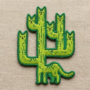 May include: An embroidered patch featuring a green cactus shape composed of five cat heads and bodies. The design uses dark green thread for the cat facial features and body stripes, with a bright green base color for the entire cactus structure.
