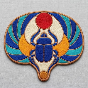 May include: An embroidered patch featuring a central blue scarab beetle topped with a red circular sun disk. The design includes symmetrical wings in shades of cream, yellow, teal, and blue, all outlined with a brown border on a fabric base.