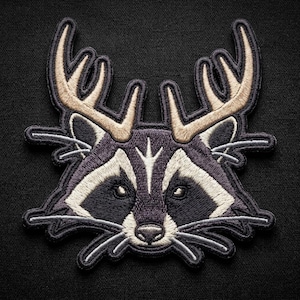 May include: An embroidered patch featuring the head of a raccoon with large, light tan antlers extending from its forehead. The raccoon has dark gray fur, white facial markings around its eyes, a black nose, and long, thin whiskers on both sides.