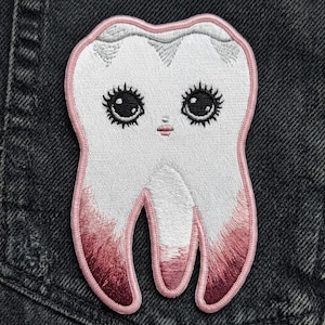 May include: An embroidered patch shaped like a tooth with a white top and pinkish-red roots. The tooth features a face with large black eyes, long eyelashes, and small pink lips, all outlined with a light pink border on a dark denim fabric.