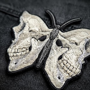 May include: An embroidered patch featuring a butterfly design where the wings are formed by two human skulls. The embroidery uses light beige and white thread for the skull details against a dark background, with a black stitched border.